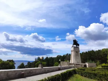 Tatishchev Monument on a tall stone pedestal beside a riverside path and low wall, with trees and water in the background.