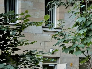 Light-colored guard house with barred windows, partly hidden by dense green foliage and an open doorway.