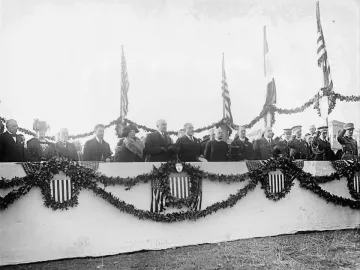 Dignitaries gathered on an outdoor platform decorated with garlands and shield emblems, with flags behind them and no statue visible.
