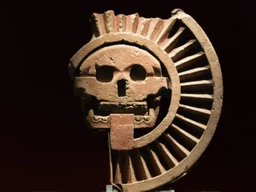 Disk of Mictlāntēcutli showing a skull-like face carved in reddish-brown stone with a spiky crown and ribbed fan-like elements.