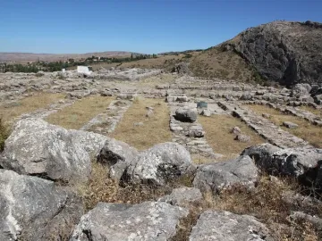 A large green stone block in front of Hattusa archaeological ruins with grassy foundations under an overcast sky.