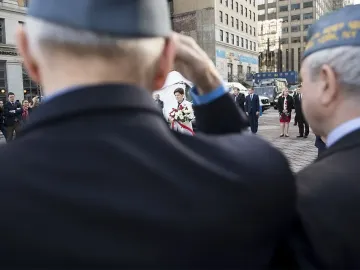 People gathered near the Katyń Memorial in an urban plaza, with one person saluting in the foreground and a woman holding white flowers in the distance.