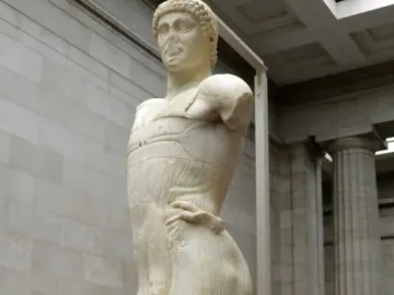 The Motya Charioteer standing on a red platform in a museum hall with columns and other artifacts nearby.