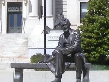Seated Lincoln sculpture of a bearded man in a suit beside a stone courthouse with columns and steps.