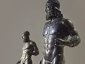 The Riace Bronzes displayed as two dark bronze male statues on light pedestals, one bearded figure in front and a smaller figure behind.