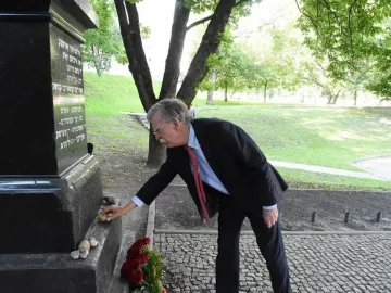 A man in a suit bends down to place a small stone at The Pit memorial, with red flowers and other stones at the base.