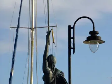 Verity sculpture seen from behind against a blue sky, framed by sailboat rigging and a lamppost.
