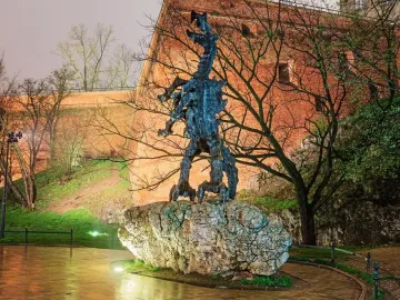 The Wawel Dragon sculpture stands on a large rock base beside a warmly lit brick wall and bare trees, with wet ground in the foreground.
