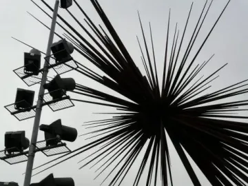 B of the Bang sculpture silhouetted against a grey sky beside a tall floodlight pole.