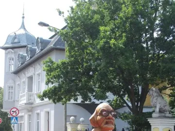 Citizen Włapko sculpture showing a naked figure with an adult head, glasses, and a beard standing on a red draped platform.