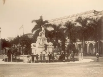 The Fuente de la India fountain in Havana with a seated female statue, adults and children gathered around it, and a large building and palm trees in the background.