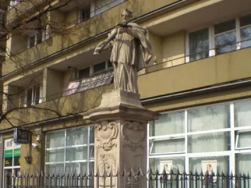 Stone statue of John of Nepomuk on an ornate pedestal behind a black spiked fence.