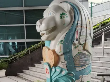 A painted Merlion sculpture with Singapore landmarks and palm trees, standing on a wave-shaped base near a staircase.