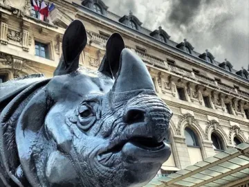 Dark rhinoceros sculpture seen from below in front of an ornate building with flags and a glass canopy.