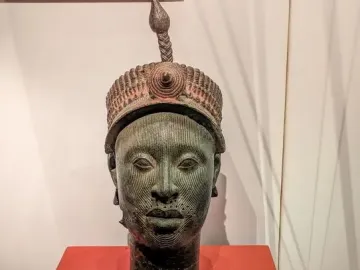 Bronze Head from Ife with green patina and intricate facial markings on a reddish pedestal.