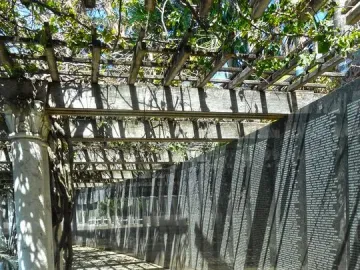 Shaded walkway beside the Holocaust Memorial’s engraved wall, with a vine-covered trellis casting patterned shadows.