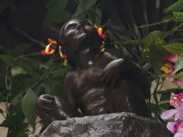 Jeanneke Pis bronze statue of a squatting young girl with ribbons in her hair, surrounded by green leaves and flowers.