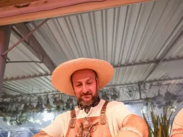 A bearded man wearing a straw hat and patterned bandana handles grilled food with tongs beside a Laçador graphic on his leather apron.