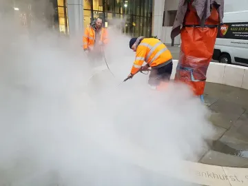 Workers clean the Pankhurst Memorial pavement with a high-pressure washer beside a wrapped vertical structure.