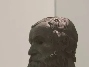 Dark patinated bronze head of the Porticello Philosopher in profile, with curly beard and hair on a light pedestal.