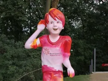 Schängel statue of a smiling boy with one hand raised to his ear, painted in red, pink, and yellow patterns.