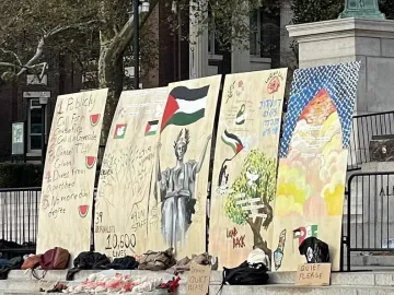 Lower portion of Alma Mater is partially visible behind protest signs and a metal barrier, with the base and part of the name visible.
