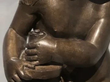 Close-up of a bronze Candelabra Angels figure with bare torso and arms, hands clasping a cylindrical object on a shallow disc.