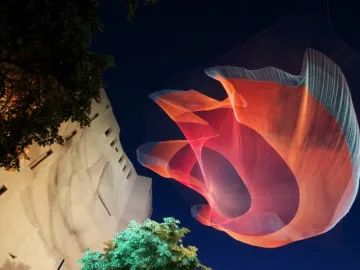 Janet Echelman’s 1.26 glows as a red and blue mesh sculpture suspended above trees at night.