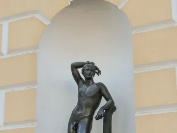 Apollino bronze nude male statue with one arm raised, standing in an arched niche beside a light yellow facade.
