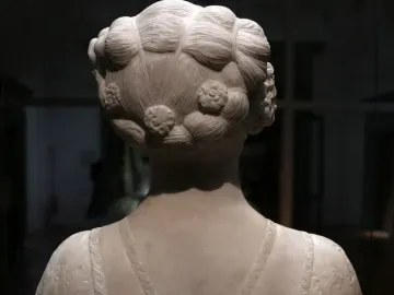 The back of a marble bust of a young woman with an elaborate coiled hairstyle and decorated drapery over the shoulders.