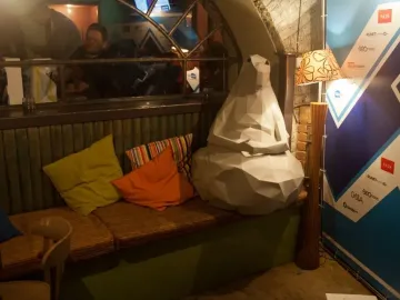 Homunculus Loxodontus sculpture seated on a padded bench among colorful pillows.
