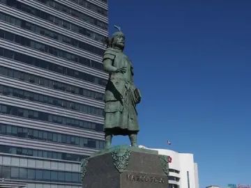 Marco Polo statue in historical attire holding a book on a stone pedestal.