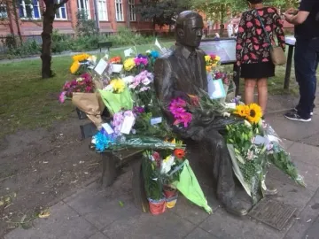 Alan Turing Memorial statue on a park bench surrounded by colorful flower bouquets.