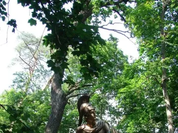 Greek Warrior Monument showing a bronze-toned armored figure with a helmet, sword, and shield among trees.