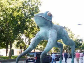 Lucky Frog sculpture with a turquoise-green patina and golden eyes on a sidewalk.