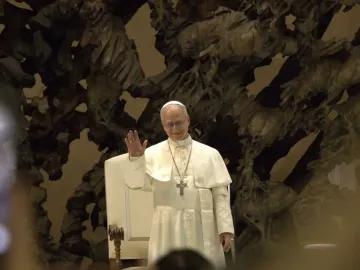 Pope Leo XIV in white clerical vestments stands in front of The Resurrection sculpture, smiling and waving.