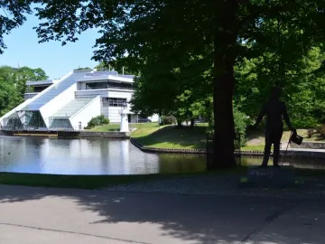 Pushkin statue on a pedestal beneath a large tree, with water and a modern building in the background.
