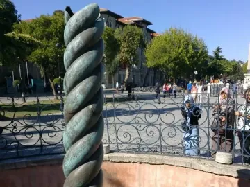 The Serpent Column with a weathered green patina stands inside a shallow red-walled enclosure near an ornate fence and public square.