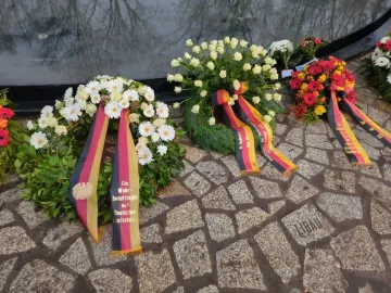 Floral wreaths with German tricolor ribbons rest on the cobblestones at the Memorial to the Murdered Sinti and Roma of Europe, beside a reflective water basin marked with the name Libau.