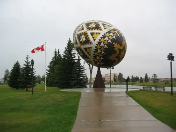 Vegreville Pysanka egg sculpture with geometric gold, black, and silver patterns in a park with trees and a pond nearby.