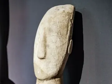 Kykladic Idol marble figurine shown from the upper chest up, with a long neck and distinct nose against a dark background.