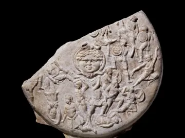 Stone relief fragment showing a central face with serpentine hair surrounded by densely packed warriors in battle.