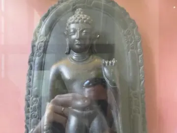 Sultanganj Buddha displayed in a glass case with a kneeling figure beside it.