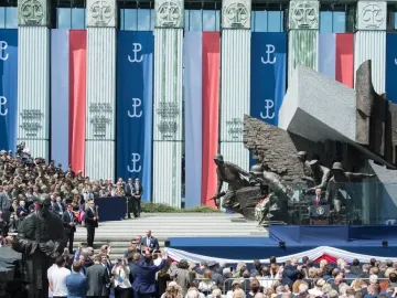 Warsaw Uprising Monument with a speaker addressing a crowd in front of it and Polish banners on a nearby building.