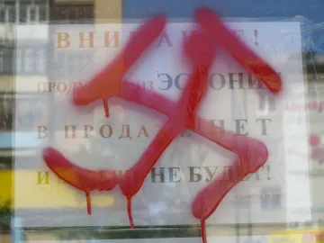 A red swastika spray-painted over a white Russian sign behind reflective glass.