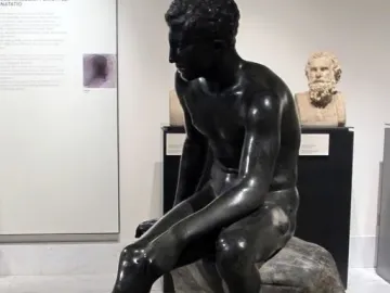 Seated Hermes statue of a nude male figure with winged sandals, shown on a rocky base in a museum display.