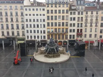 Fontaine Bartholdi in the center of Place des Terreaux, surrounded by wet paving, pedestrians, and surrounding buildings.