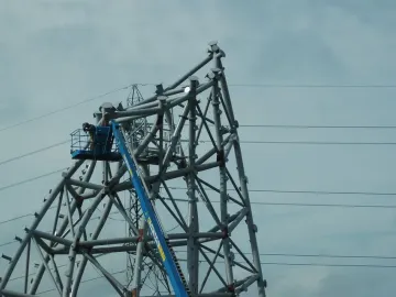 A large grey metal sculpture under construction with a worker in a blue boom lift nearby and power lines overhead.