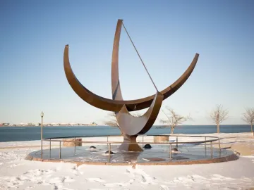 Man Enters the Cosmos sundial sculpture on a circular base beside a snowy lakeshore, with a city skyline in the distance.