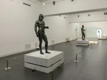 The Riace bronzes displayed on white pedestals in a brightly lit museum hall.